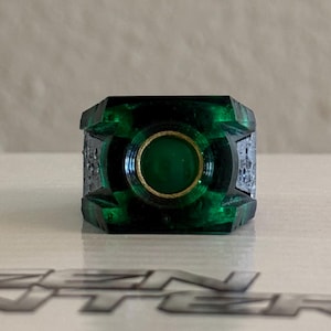 Peut inclure: Une bague vert foncé avec un motif circulaire central et un centre doré. La bague a un design géométrique et est posée sur une surface blanche.