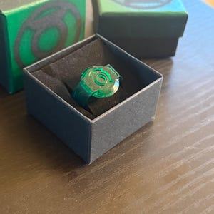 May include: A translucent green ring with a circular emblem, displayed in a black box. The ring is a vibrant green colour, and the box is inside a larger green box with a matching emblem. The ring is a collectible item.