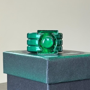 May include: A green metallic ring with the Green Lantern symbol. The ring has a raised, circular design with a central green gem. It is displayed on a dark green box, highlighting the ring's colour and design.
