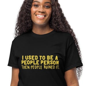 Puede incluir: Camiseta negra con el texto amarillo "I USED TO BE A PEOPLE PERSON THEN PEOPLE RUINED IT". La camiseta la lleva una persona con pelo largo y rizado oscuro y pantalones color beige.