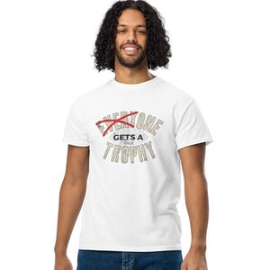 Może przedstawiać: Biała koszulka z napisem "Everyone Gets a Trophy" w stylu vintage. Słowo "Everyone" jest przekreślone na czerwono. Osoba ma na sobie niebieskie dżinsy i białe trampki.