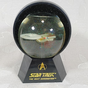 May include: A Star Trek The Next Generation snow globe featuring a miniature USS Enterprise starship inside a clear sphere filled with iridescent glitter. The base is black with a gold Starfleet insignia and the text Star Trek The Next Generation.