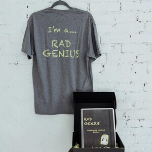 Könnte beinhalten: Ein graues T-Shirt mit dem handgeschriebenen Text "I'm a.... RAD GENIUS" hängt an einem Kleiderbügel. Darunter befindet sich eine schwarze Box mit einem Notizbuch mit "RAD GENIUS" auf dem Cover. Das Notizbuch ist die Radiologie-Assistenten-Ausgabe.