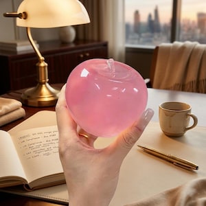 Peut inclure: Une sculpture de pomme en verre rose translucide et lisse avec une tige en verre transparent. L'objet est sphérique avec une finition brillante, reflétant la lumière sur sa surface, et est tenu dans une main sur un bureau avec une lampe.