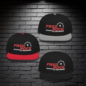 Könnte beinhalten: Drei schwarze Baseballkappen mit dem Text "FRED ROCK SCHOOL OF DRUMMING" in Rot und Weiß. Jede Kappe hat einen unterschiedlich farbigen Schirm: rot, grau und schwarz. Die Kappen werden vor einer dunklen Backsteinmauer präsentiert.