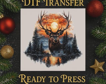 Op de afbeelding: Een DTF-transfer met een majestueuze hert met een groot gewei, tegen een achtergrond van een zonsondergang boven een bos en meer. De tekst "DTF TRANSFER" en "READY TO PRESS" is goudkleurig. De afbeelding is omgeven door kerstversieringen.