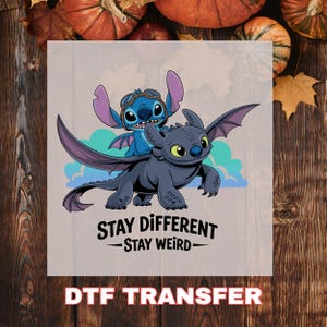 May include: DTF transfer featuring Stitch and Toothless from "How to Train Your Dragon" and "Lilo & Stitch". The image shows Stitch wearing goggles and Toothless with outstretched wings. The text reads "Stay Different - Stay Weird".
