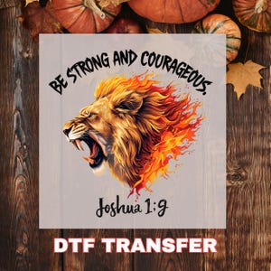 May include: A DTF transfer featuring a roaring lion with a fiery mane and the text "Be Strong and Courageous" above and "Joshua 1:9" below. The lion is rendered in shades of gold, orange, and red.