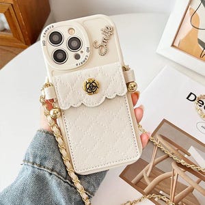 Puede incluir: Funda de teléfono de color crema con textura acolchada y un pequeño bolsillo. Cuenta con una correa de cadena dorada y una flor dorada decorativa. El área de la cámara tiene las palabras "HD camera".