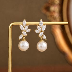 May include: A pair of gold-tone drop earrings featuring a cluster of marquise-cut clear cubic zirconia stones arranged in a leaf-like pattern, with a single round white pearl suspended from the base of each earring on a gold-tone metal post.