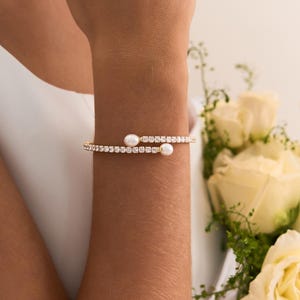 May include: A gold-toned wrap bracelet featuring a continuous row of clear, sparkling crystals and two small, round white pearls at each terminal end. The flexible design allows the bracelet to coil around the wrist for a secure fit.