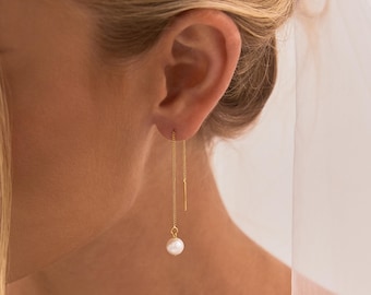 May include: A gold threader earring featuring a delicate chain that passes through the earlobe, ending in a single round white pearl drop. The earring is worn in the ear, with the chain hanging down and the pearl suspended at the bottom.