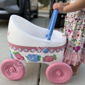 May include: A white toy stroller with pink wheels and blue handles. The stroller is decorated with pink and blue floral designs and a pink trim. The wheels are a solid pink color.