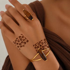 May include: A gold-toned ring and a matching gold-toned cuff bracelet, both featuring a smooth, elongated, dark brown oval stone. The jewelry set is displayed alongside two square-shaped temporary tattoos with a brown leopard print pattern.