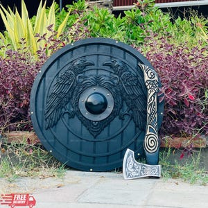 May include: A circular black shield featuring a central embossed bear head flanked by two ravens, with a matching black and silver axe leaning against it, decorated with intricate carved patterns on the handle and the metal blade.