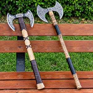 May include: Two double-bit axes with silver-colored metal heads featuring intricate etched patterns. The left axe has a light wood handle with dark leather wrapping. The right axe has a carved wood handle with black leather grip sections.