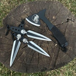 May include: A black tomahawk axe and three throwing knives with skull designs. The axe and knives have silver blades and black handles. The items are displayed on a wooden surface.