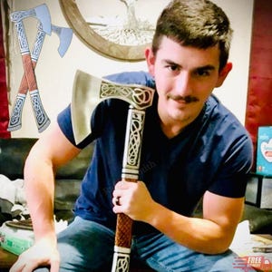 May include: Axe with a silver blade and a decorative handle. The handle has a brown, textured grip and is adorned with intricate white patterns. The axe head features a detailed design. The axe is held by a person wearing a blue shirt and jeans.