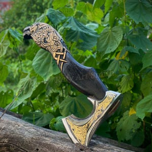 May include: A handcrafted axe with a black handle and a gold-colored blade. The handle features intricate carvings and a detailed animal head design. The blade has decorative gold patterns. The axe is displayed on a wooden log against a backdrop of green foliage.