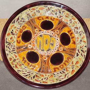 May include: A ceramic plate with a maroon rim and a central design in shades of yellow, orange, and green. The plate features a circular pattern with text in the centre and dark maroon circles. The plate is likely for decorative or ceremonial use.