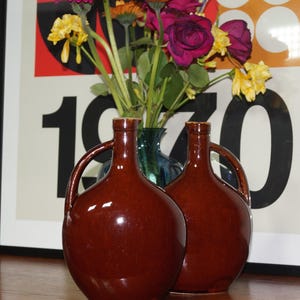 May include: Two brown ceramic jugs with handles. The jugs are in front of a vase of flowers and a framed poster with the number 1970. The jugs have a glossy finish and a rounded shape.