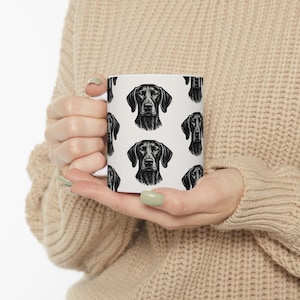 May include: White ceramic mug featuring a repeating pattern of a black dog's head. The dog's portrait is detailed, with a focus on the eyes and snout. The mug is held in a person's hands, with a neutral background.