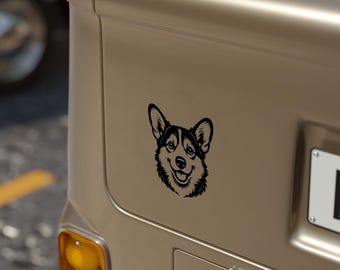 May include: A black and white corgi dog sticker is attached to the back of a vehicle. The sticker shows a smiling corgi head with detailed fur and facial features. The vehicle is a light tan colour.