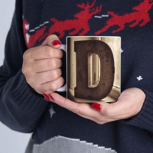 May include: White ceramic mug featuring a large, distressed brown letter "D" against a sepia-toned background. The mug has a standard handle and a glossy finish. The mug is held by a person wearing a navy blue sweater.