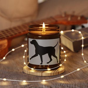 May include: A lit brown glass candle jar featuring a black dog silhouette on a white label. The candle is on a wooden coaster, with a string of small lights around it. A guitar and a book are in the background.