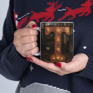 May include: White ceramic mug featuring a close-up of a rusty metal letter "T" illuminated with small lights. The mug is held in hands, and the background is blurred.