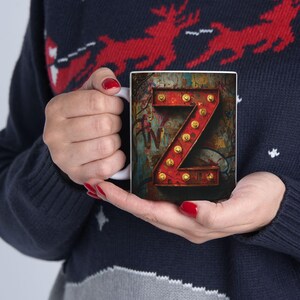May include: White ceramic mug featuring a red letter "Z" illuminated with golden light bulbs. The letter is set against a colorful, distressed background with graffiti-style art. The mug is held by a person wearing a blue sweater.