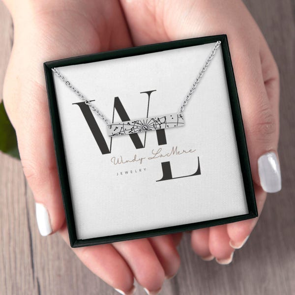 May include: A silver necklace with a rectangular pendant featuring a compass design, presented in a black jewelry box. The box has the Windy LaMere Jewelry logo on the inside. The necklace is held by a person.