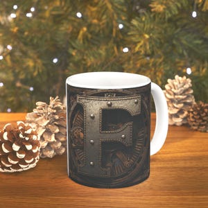 May include: White ceramic mug featuring a detailed, steampunk-style letter "F" in shades of gray and brown. The design incorporates gears and rivets, set against a dark background. The mug has a white handle and is placed on a wooden surface.