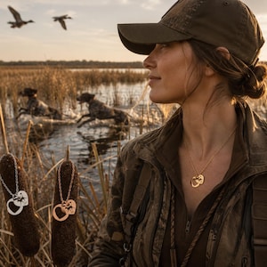 May include: A woman wearing a camouflage jacket and cap, with a gold necklace featuring a heart and bird pendant. Two similar necklaces hang from cattails in the foreground. Two dogs run through the water in the background.