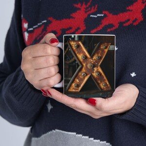 May include: White ceramic mug featuring a close-up of a rusty, vintage letter "X" with light bulbs. The "X" is brown and gold, with visible rust and wear. The mug is held by a person wearing a dark blue sweater.
