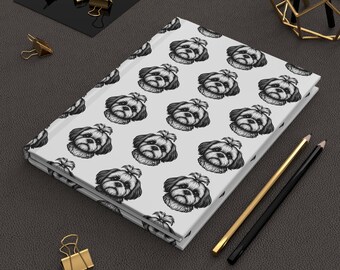 May include: A white journal featuring a repeating pattern of a Shih Tzu dog illustration. The journal is rectangular and closed, with a glossy cover. Gold and black pencils and paper clips are arranged around the journal on a dark gray surface.