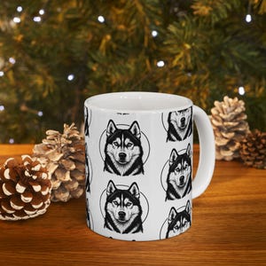 May include: White ceramic mug featuring a repeating pattern of a black and white husky dog portrait within a circle. The mug has a white handle and is set against a blurred background with pine cones.