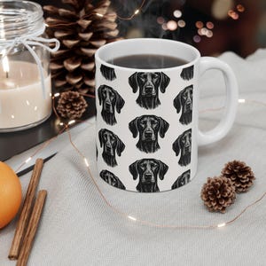 May include: White ceramic coffee mug featuring a repeating pattern of black dog portraits. The mug is filled with a dark liquid and has a white handle. The design is a black and white illustration of a dog's head.