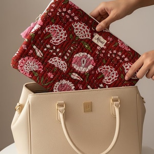 Può includere: Una borsa color crema con dettagli dorati. Una custodia per laptop trapuntata con motivo floreale rosso viene inserita all'interno. La custodia ha un nappa rosa e una cerniera bianca. Il motivo floreale comprende fiori rosa e bianchi.