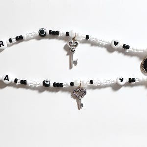 Puede incluir: Dos pulseras de cuentas blancas y negras con abalorios. Una pulsera tiene la letra "R", un dije de llave y un dije de luna. La otra pulsera tiene la letra "A", un dije de llave y un dije de luna.