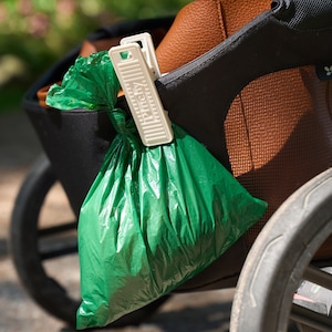 May include: A green plastic waste bag is clipped to a stroller with a white clip. The clip has the word "Puchy" printed on it. The stroller has a black frame and brown leather accents.