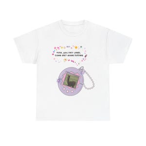 Könnte beinhalten: Weißes T-Shirt mit einem lila Digital-Haustier-Spielzeug-Design mit einem Lama-Bild. Der Text über dem Spielzeug lautet "TINA, YOU FAT LARD, COME GET SOME DINNER". Das Design enthält dekorative Sterne und Herzen.