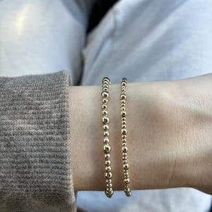 May include: Two gold-toned beaded bracelets worn together. One bracelet features a mix of small and medium-sized round beads, while the other consists of uniform, smaller round beads. Both bracelets have a polished, reflective metallic finish.