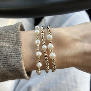 May include: Three stretch bracelets featuring a combination of round white pearls and small metallic gold-toned beads. The bracelets are arranged in a stack, alternating between pearl-heavy designs and a design composed primarily of gold beads.