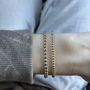 May include: Two gold-toned beaded bracelets worn together on a wrist. Each bracelet consists of a single strand of small, uniform, polished spherical beads that create a continuous circular design with a reflective metallic finish.