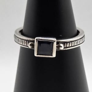 Peut inclure: Bague en argent avec une pierre noire carrée au centre. L'anneau présente un motif texturé détaillé avec des lignes verticales et de petits accents circulaires. La bague est présentée sur un support noir.
