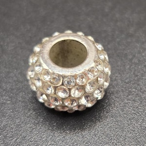 May include: A silver-colored bead with a spherical shape, encrusted with numerous small, clear crystals. The bead has a central hole running through it. The background is a dark gray color.