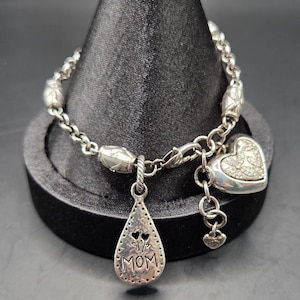 May include: Silver bracelet with a chain-link design, featuring decorative beads and charms. Charms include a heart with embedded crystals and a teardrop pendant with the word "MOM". The bracelet is displayed on a black stand.
