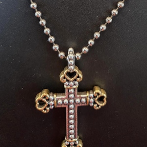 以下が含まれることがあります： 銀色のボールチェーンネックレスと装飾的な十字架ペンダント。十字架は、金色の透かし模様のデザインと、透明なクリスタルのアクセント、中央のクリスタルの列が特徴です。ネックレスは黒い背景に表示されています。
