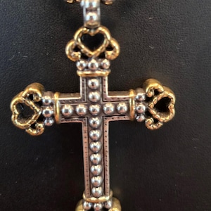 以下が含まれることがあります： ゴールドカラーのアクセントとビーズチェーンが付いた、シルバーカラーのクロス型ペンダント。クロスは、一連の小さな丸い装飾と装飾的な装飾が施された詳細なデザインが特徴です。ペンダントは暗い背景にぶら下がっています。